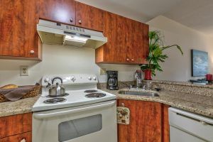 A cozy kitchen with wooden cabinets, featuring a stove, oven, coffee maker, and a teapot on the counter.