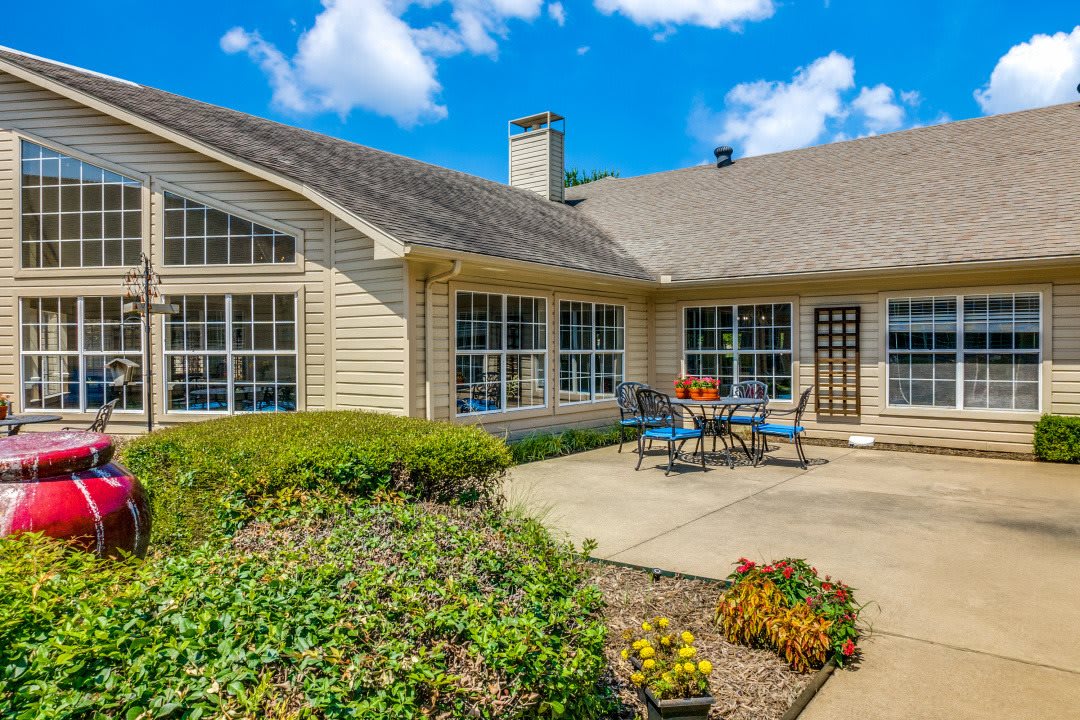A cozy patio with outdoor furniture in front of a house, surrounded by a garden on a sunny day.
