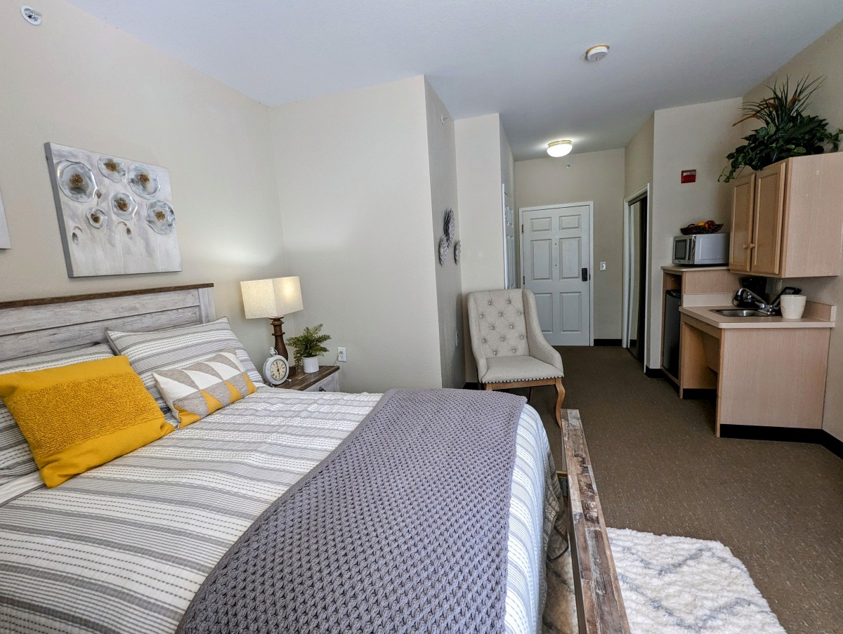 A cozy bedroom featuring a neatly made bed with decorative pillows, a nightstand with a lamp, and a comfortable chair near the door.