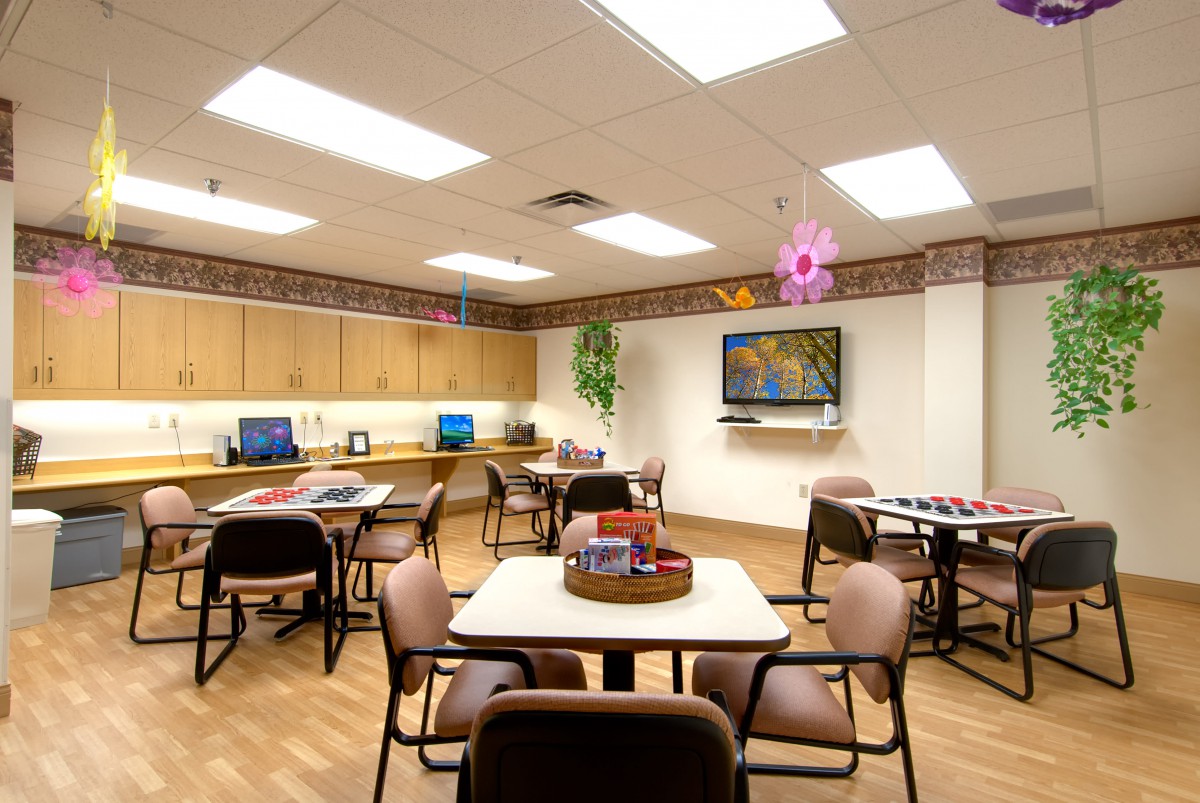 A well-lit room with multiple tables and chairs, featuring a television and computer workstations along the wall.