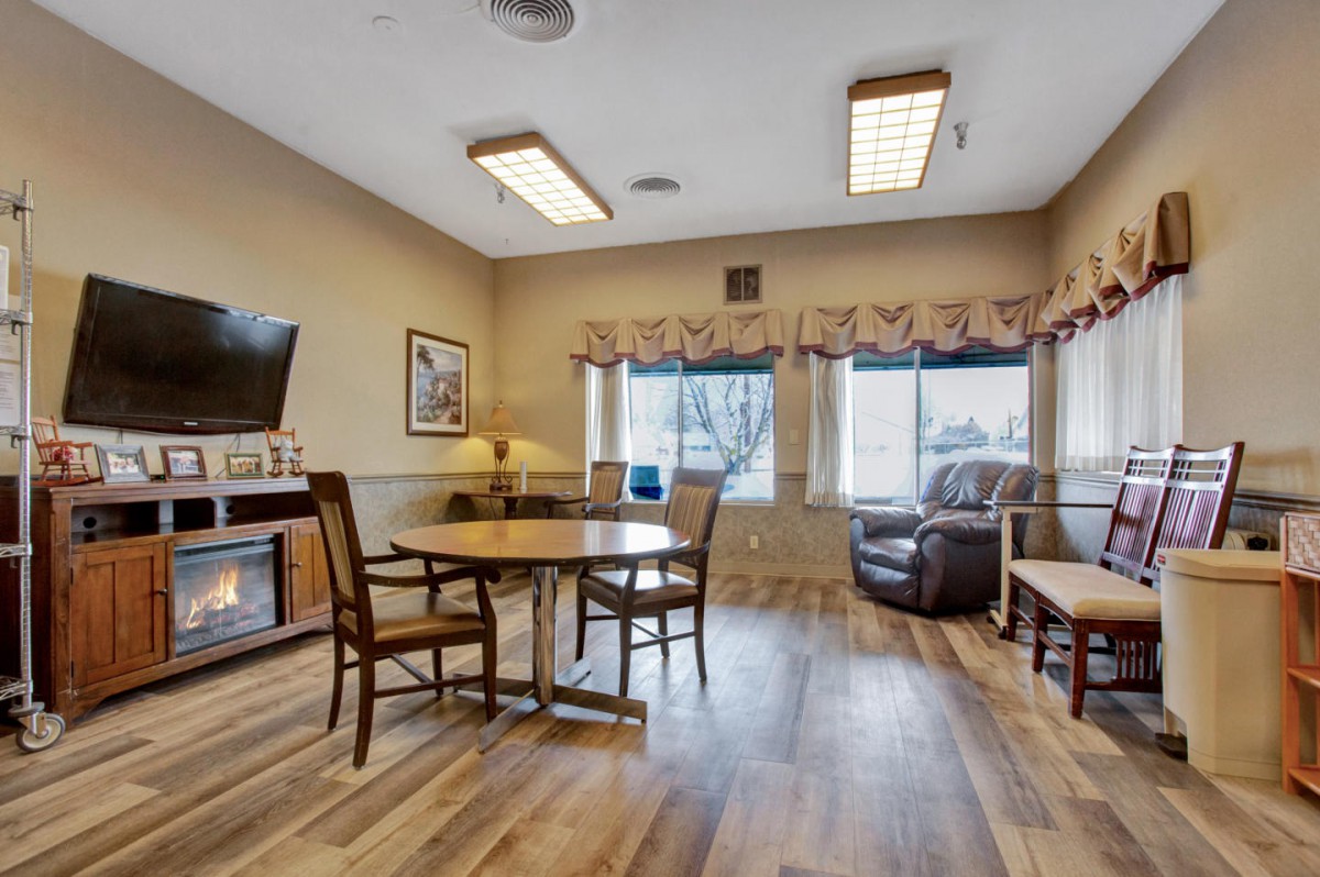A cozy living room with wooden flooring, featuring a table and chairs set, an armchair, and a television.