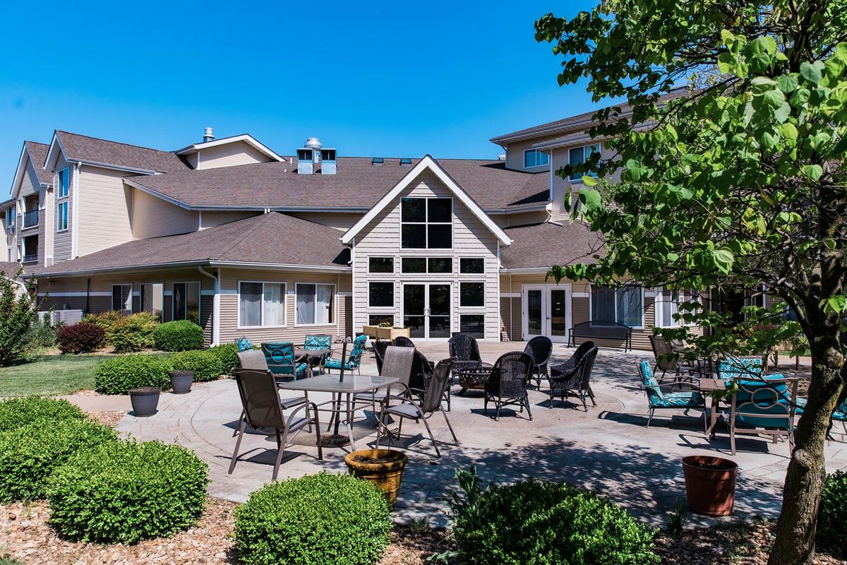 A spacious outdoor patio area with tables and chairs in front of a large building, surrounded by well-maintained landscaping under a clear blue sky.