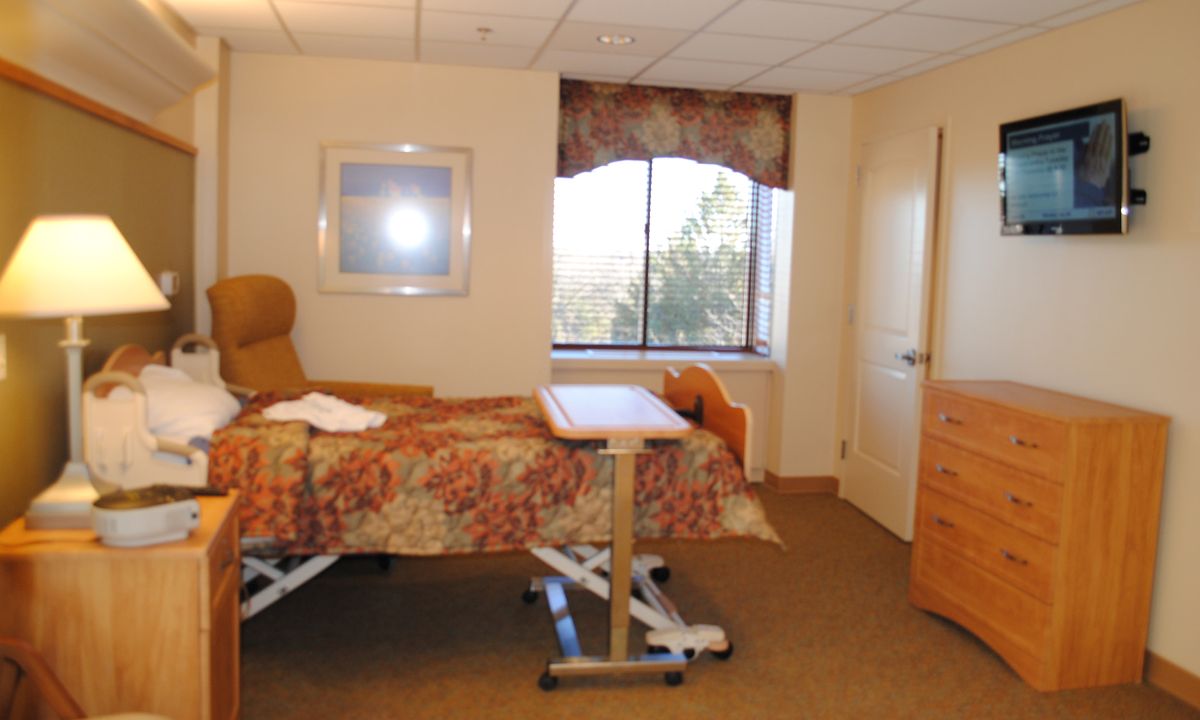 A hospital room with a patient bed and medical equipment, featuring a window and comfortable seating.
