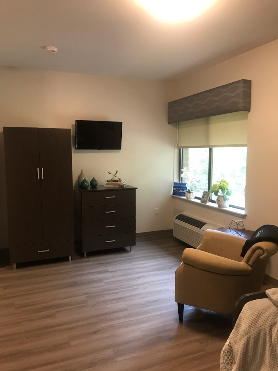 A cozy, well-furnished room featuring a wardrobe, dresser, wall-mounted TV, comfortable chair, and window with natural light.