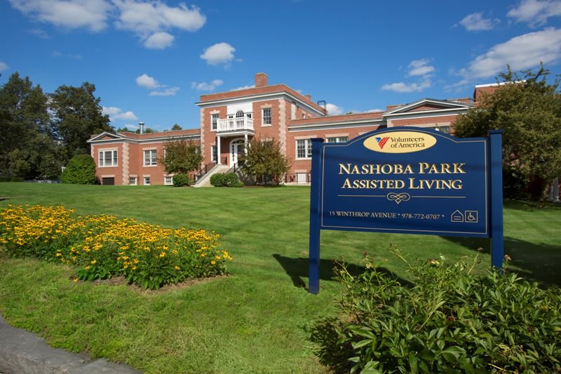 A scenic view of Nashoba Park Assisted Living with a brick building and manicured lawn.