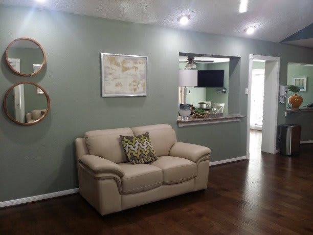 A modern living room with a beige sofa against a green wall, decorated with wall art and mirrors, featuring an open view to the kitchen area.