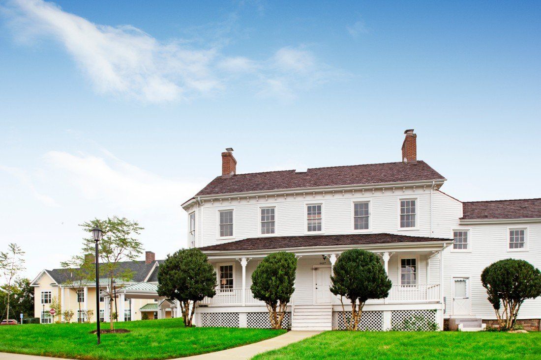 A large, two-story white house with a porch and surrounding greenery under a clear blue sky.