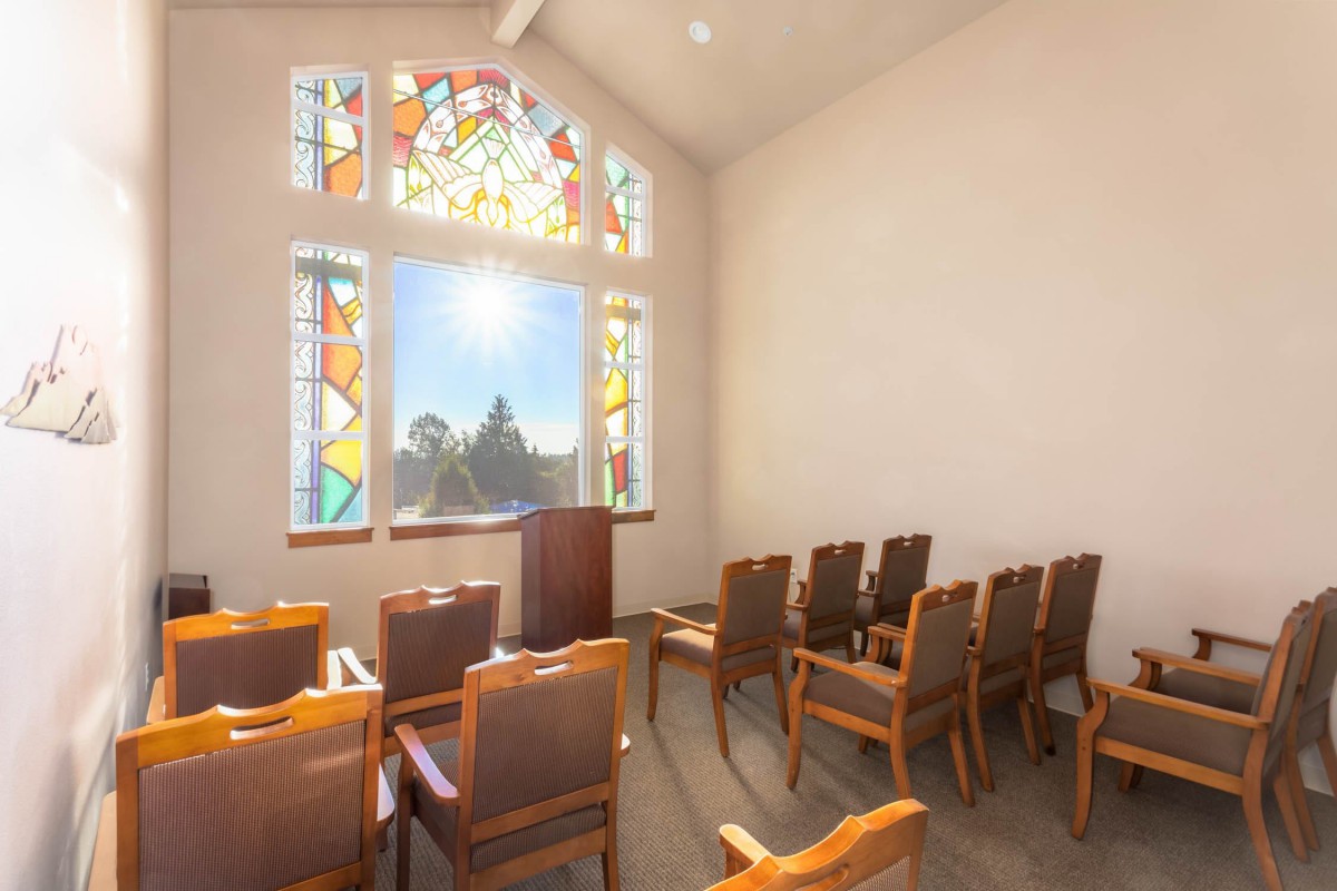 A chapel with rows of chairs facing a pulpit, illuminated by sunlight through a stained glass window.