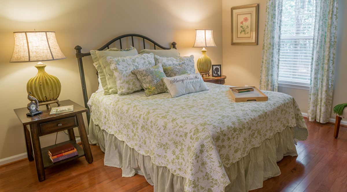 A cozy bedroom with a neatly made bed featuring floral patterned bedding, multiple pillows, and a warm lamp on the nightstand.