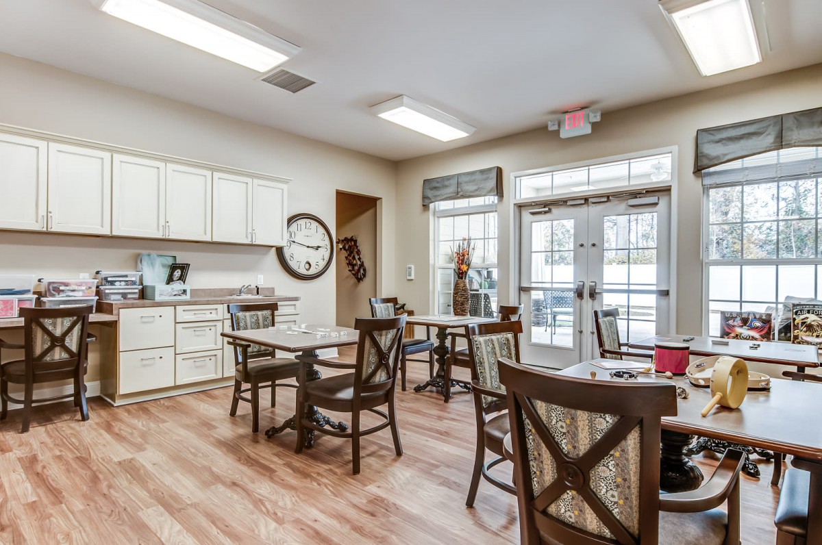 A cozy kitchen and dining area with wooden floors, featuring several tables and chairs, a clock on the wall, and bright natural lighting.