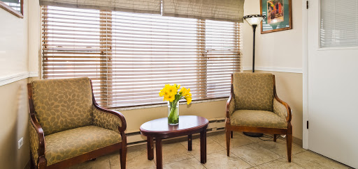 A cozy living room featuring two chairs, a small table with vibrant yellow flowers, and soft natural light filtering through the blinds.