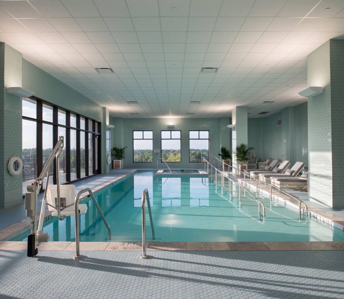 A serene indoor swimming pool with natural light streaming in through large windows, surrounded by comfortable lounge chairs.