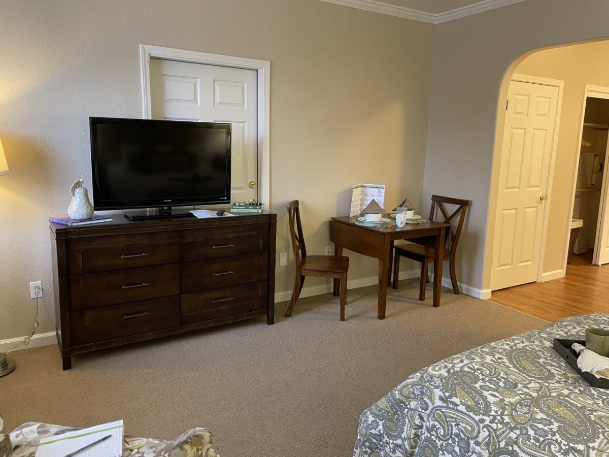 A cozy hotel room featuring a television on a dresser, a small desk with a chair, and neutral decor.
