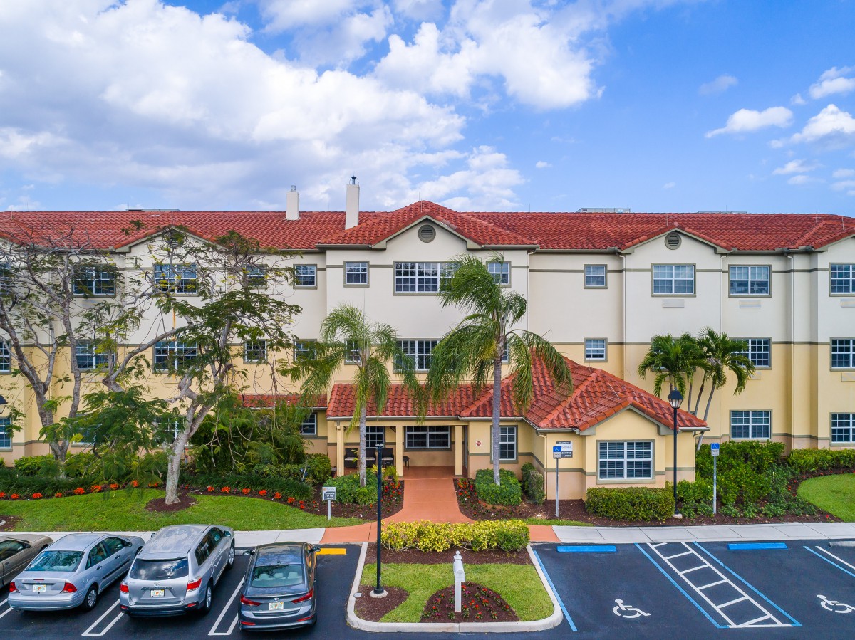 A residential apartment complex with a red-tiled roof and landscaped surroundings, featuring a parking lot in the foreground.