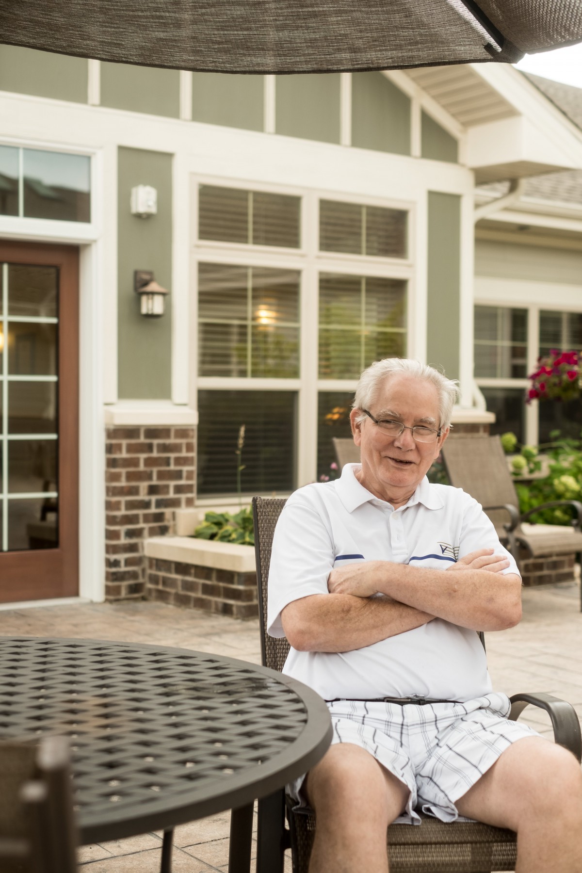 An elderly man sitting outside on a patio in front of a residential home.