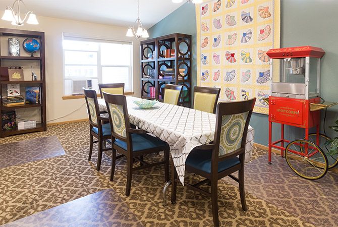 A cozy dining room with a large table, surrounded by chairs, featuring a patterned carpet and a colorful quilt wall hanging.