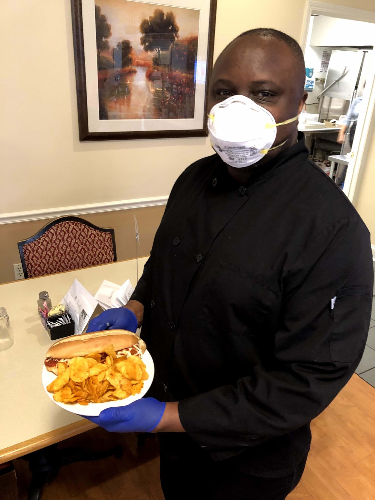 A chef wearing a mask and gloves presents a sandwich with chips in a restaurant setting.