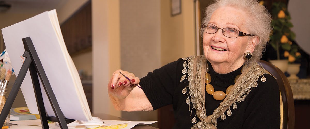 An elderly woman with glasses is smiling while sitting indoors.