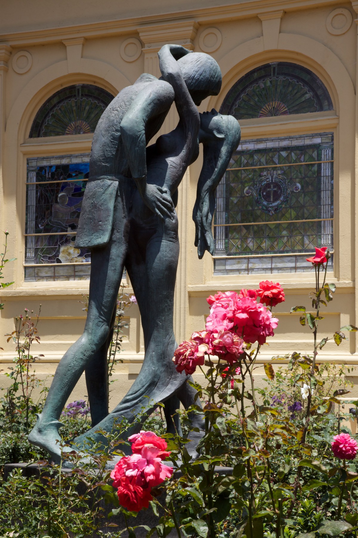 A sculpture of two figures embracing, surrounded by blooming roses in front of a building.