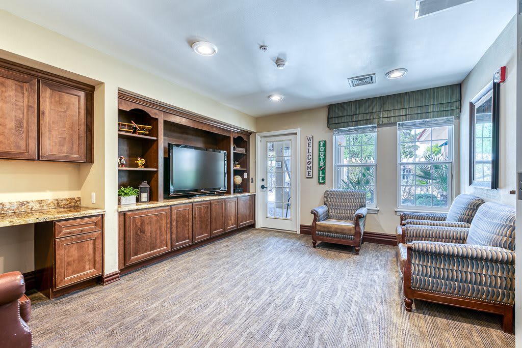 A cozy living room with wooden flooring, a television, bookshelves, and comfortable seating near large windows.