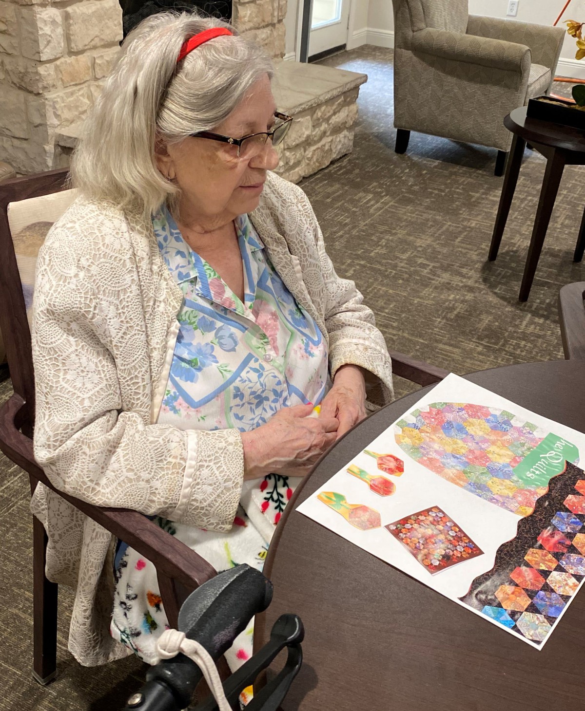 An elderly woman wearing glasses is seated at a table, observing some artwork.
