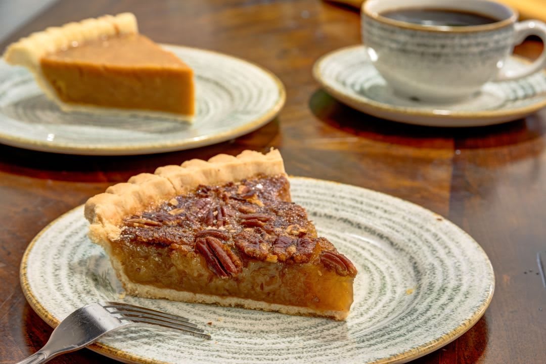 A slice of pecan pie on a plate with a cup of coffee in the background.