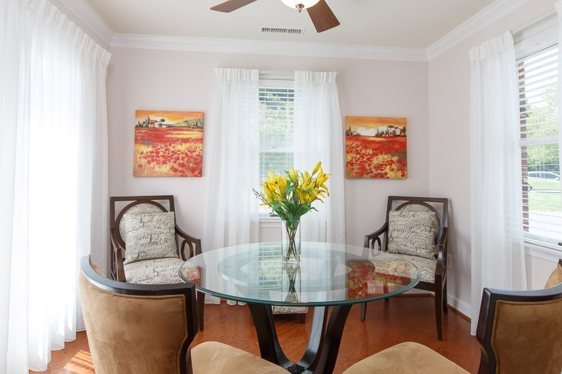 A cozy living room features a glass table surrounded by elegant chairs, with a vibrant floral arrangement and colorful artwork adding charm.