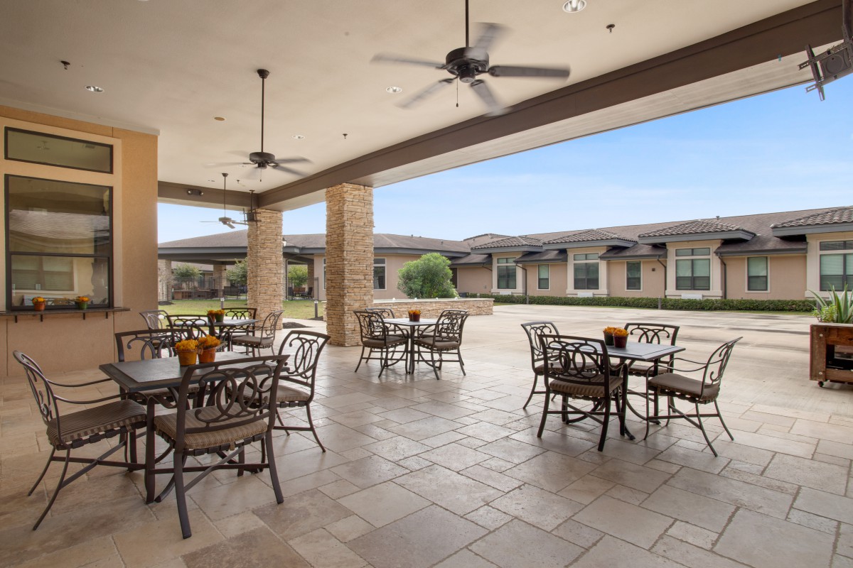An outdoor patio with tables and chairs, covered by a roof with ceiling fans, offering a view of a landscaped area.