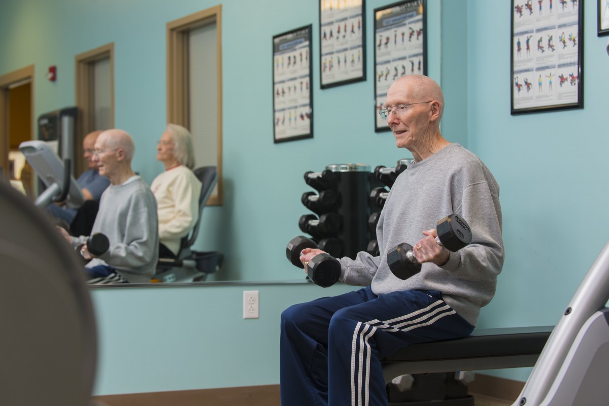 An elderly man exercises in a gym, lifting dumbbells while seated and looking at the mirror.