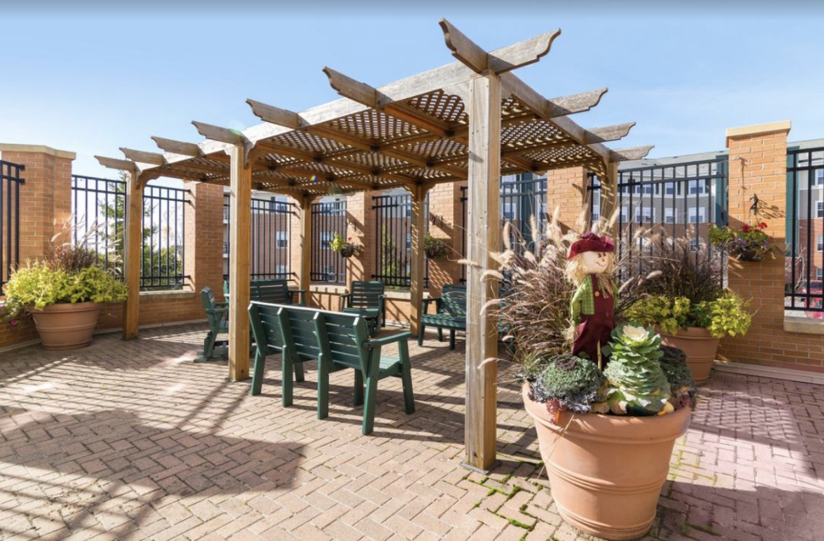 A sunny patio featuring a wooden pergola with green chairs and large planters.