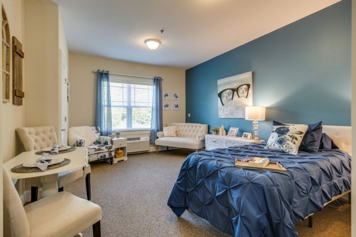 A cozy bedroom with a blue accent wall, featuring a bed, comfortable seating, and a window allowing natural light.