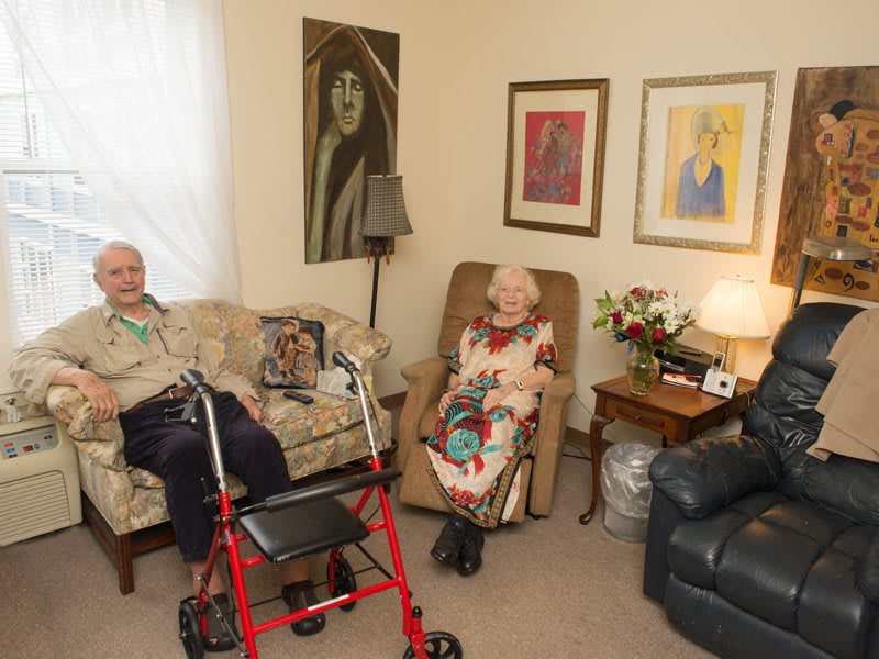 An elderly couple sitting in a cozy living room with a walker nearby and various portraits on the walls.