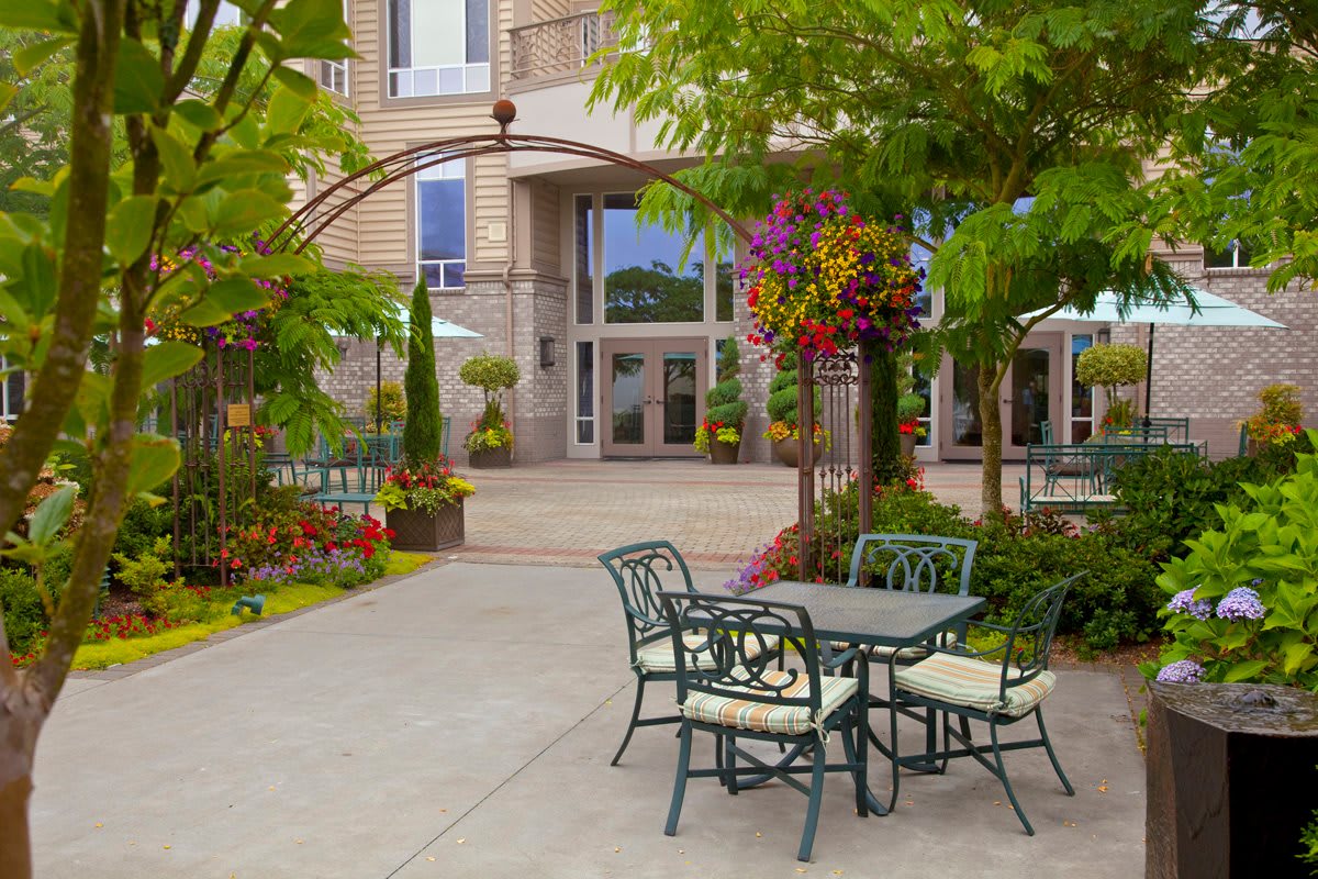 A charming outdoor patio area with a table and chairs surrounded by lush greenery and colorful flowers in front of a building.