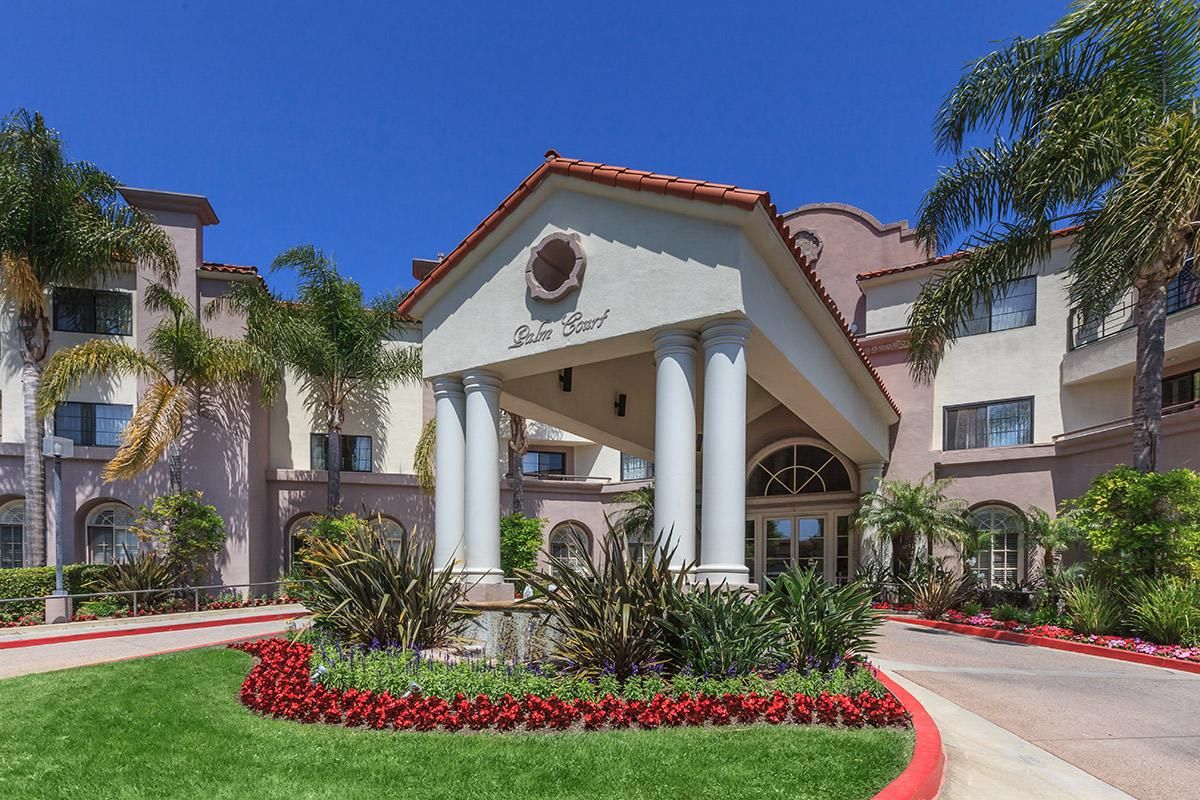 A picturesque entrance of a building featuring columns, surrounded by lush landscaping and palm trees.
