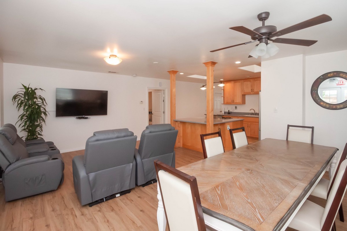 A modern open floor plan featuring a living room with recliners, a dining area with a wooden table, and a kitchen in the background.