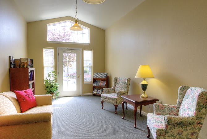 A cozy living room with large windows, featuring a sofa, chairs, and a lamp.