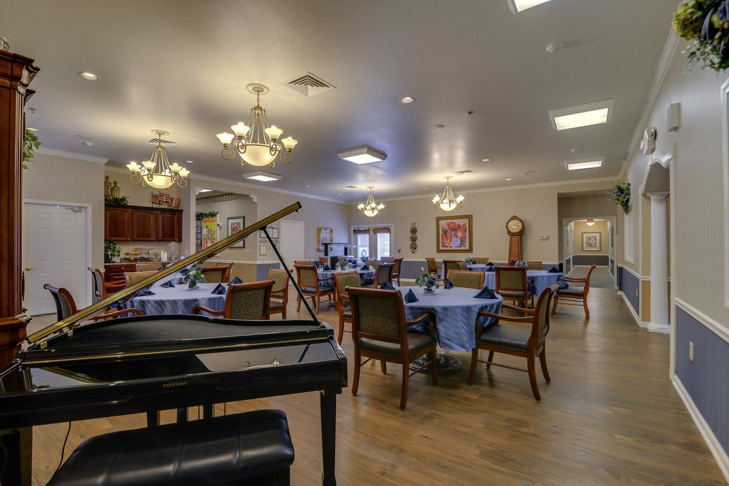 A spacious dining room featuring elegantly set tables, comfortable chairs, and a grand piano under warm chandeliers.