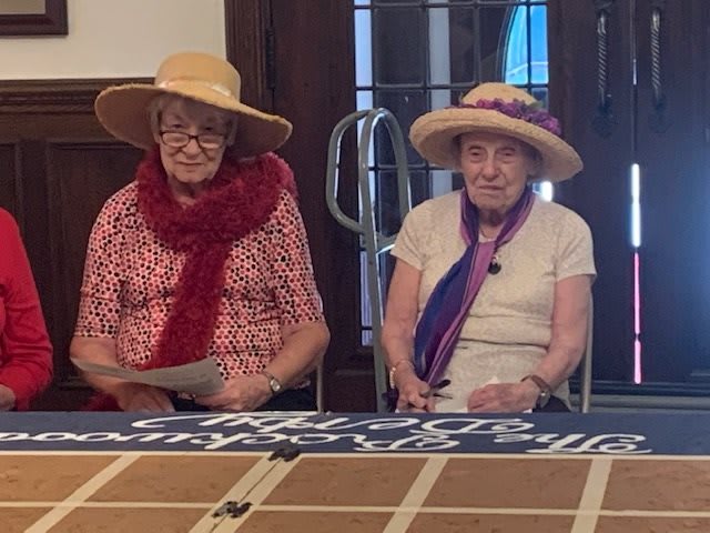 Two elderly women wearing hats and scarves sit at a table indoors.