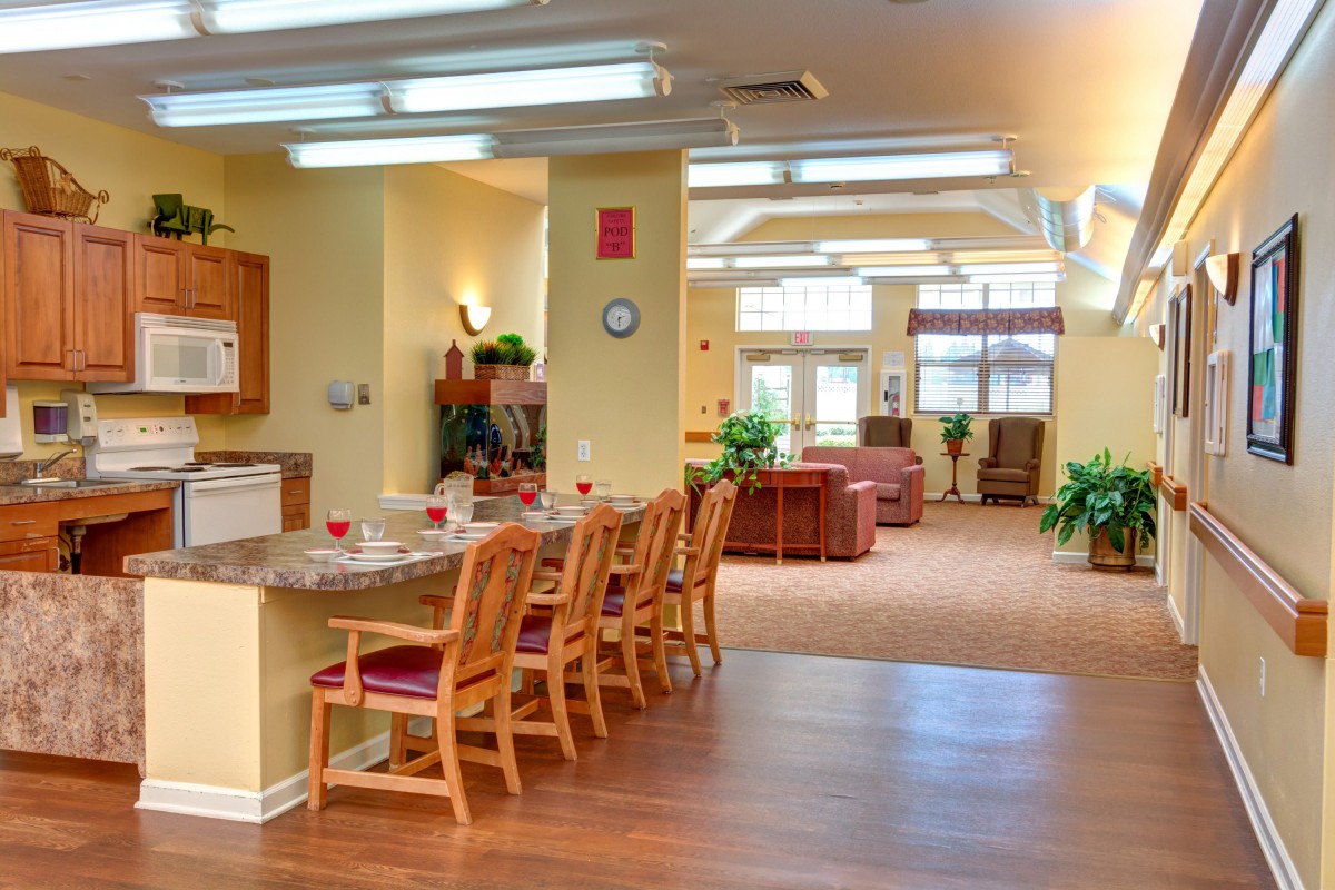 A bright kitchen and dining area with wooden chairs and tables, featuring plants and warm lighting.