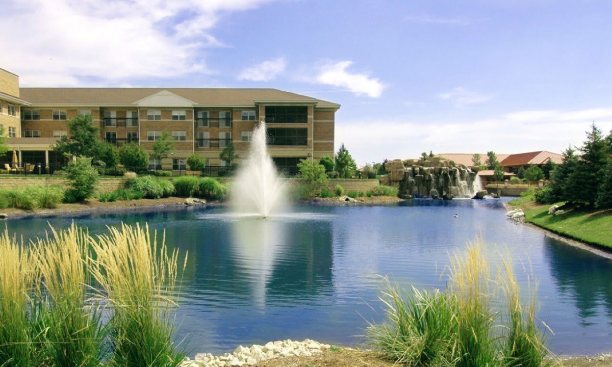 A serene pond with a fountain in front of a building, surrounded by lush greenery and a clear sky.