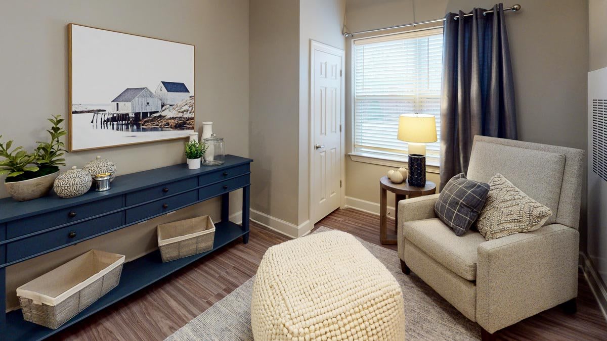 A cozy living room corner with an armchair, a pouf, and a blue cabinet, illuminated by a lamp near the window.