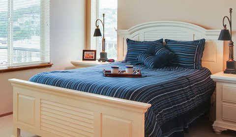 A cozy bedroom featuring a bed with blue striped bedding, accentuated by a tray with cups and a lamp beside a window.