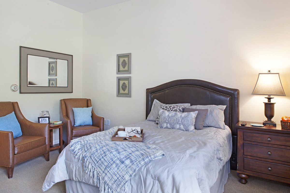 A cozy bedroom featuring a neatly made bed with decorative pillows, an armchair, a lamp, and framed art on the walls.