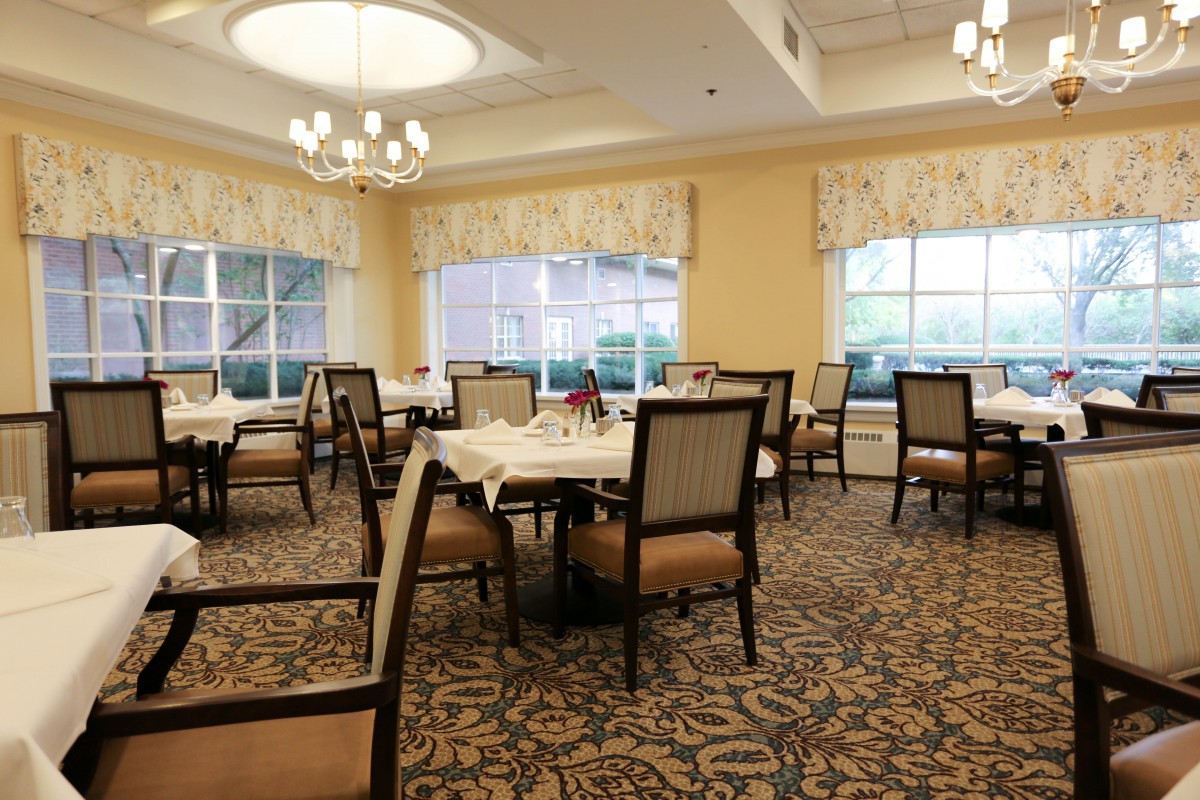 An elegant dining room with neatly arranged tables and chairs, large windows, and a chandelier.