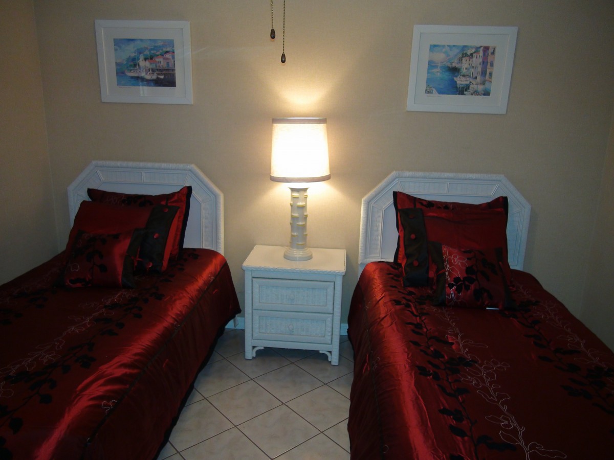A cozy bedroom with twin beds adorned in red bedding, a shared white nightstand, and soft lighting from a table lamp.