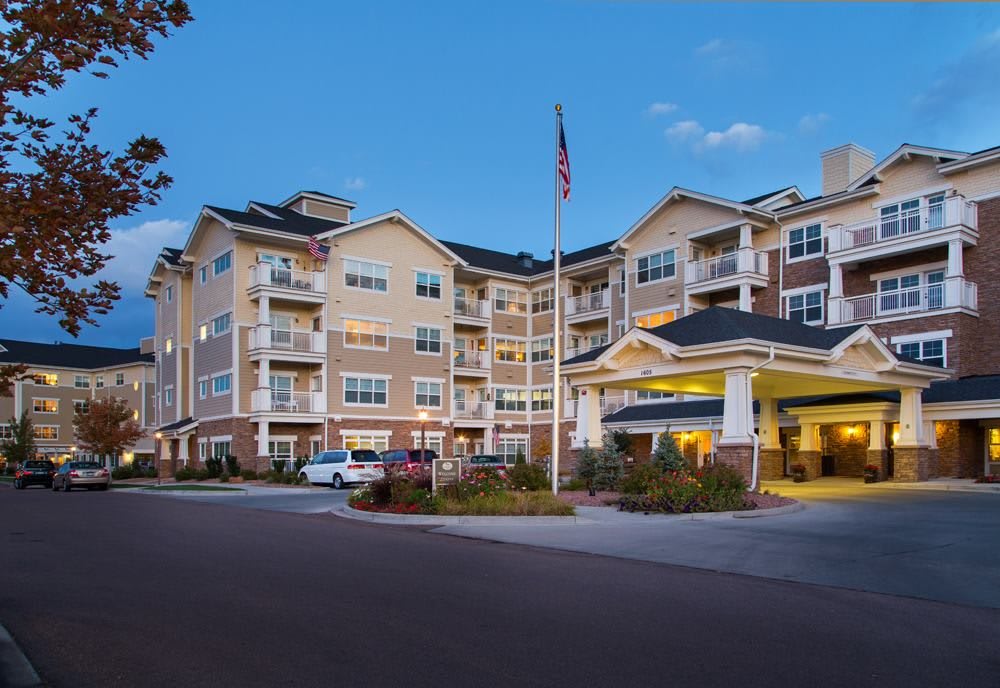 A multi-story residential building with balconies and an American flag in the foreground, set against an evening sky.