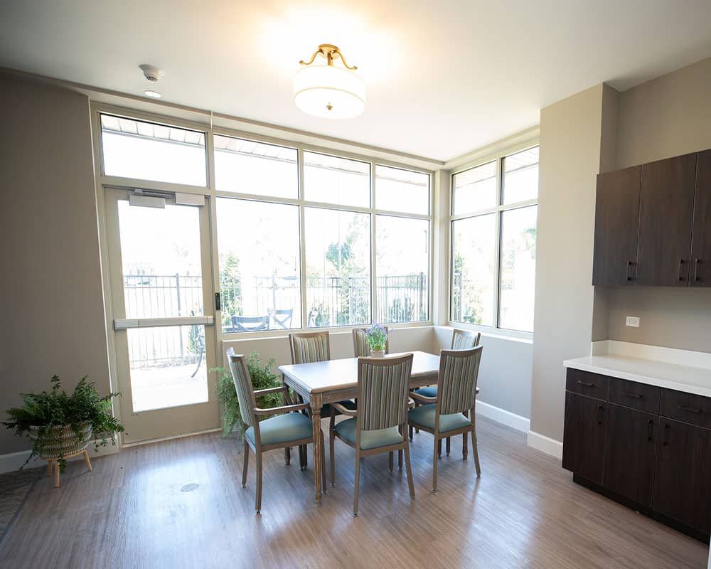 A bright dining area with large windows, a table, and chairs surrounded by natural light and indoor plants.