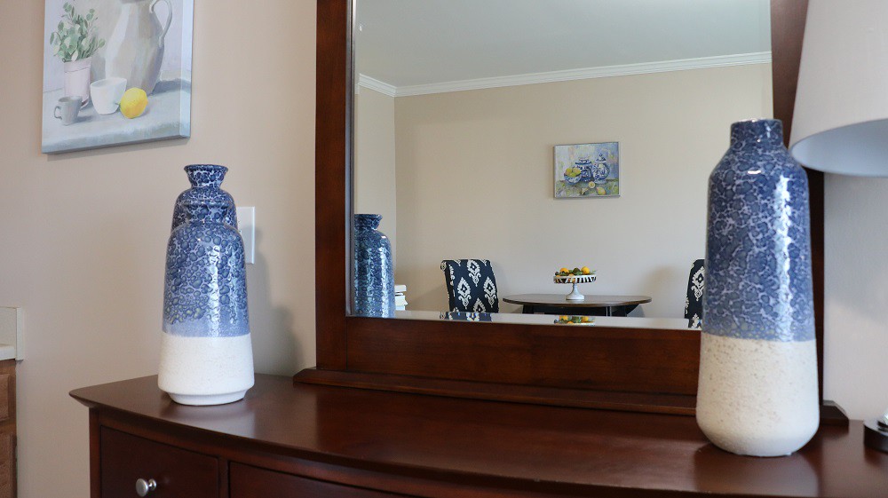 A wooden dresser with a large mirror reflecting a dining room setup, featuring a blue and white vase.