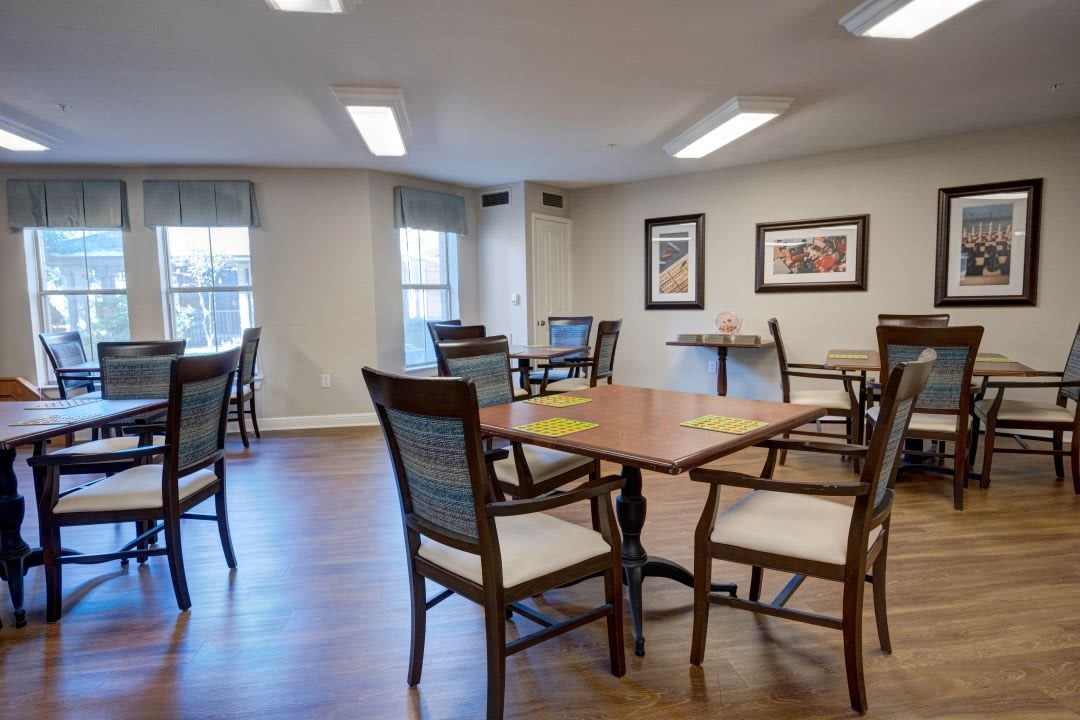 A cozy dining area with wooden tables and chairs, featuring artwork on the walls and natural light from large windows.