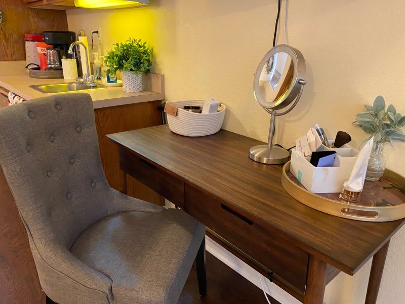 A cozy makeup station with a wooden desk, plush chair, and various accessories.